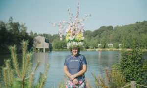 Jule Harries wearing a flowery hat, stood in front of a lake.